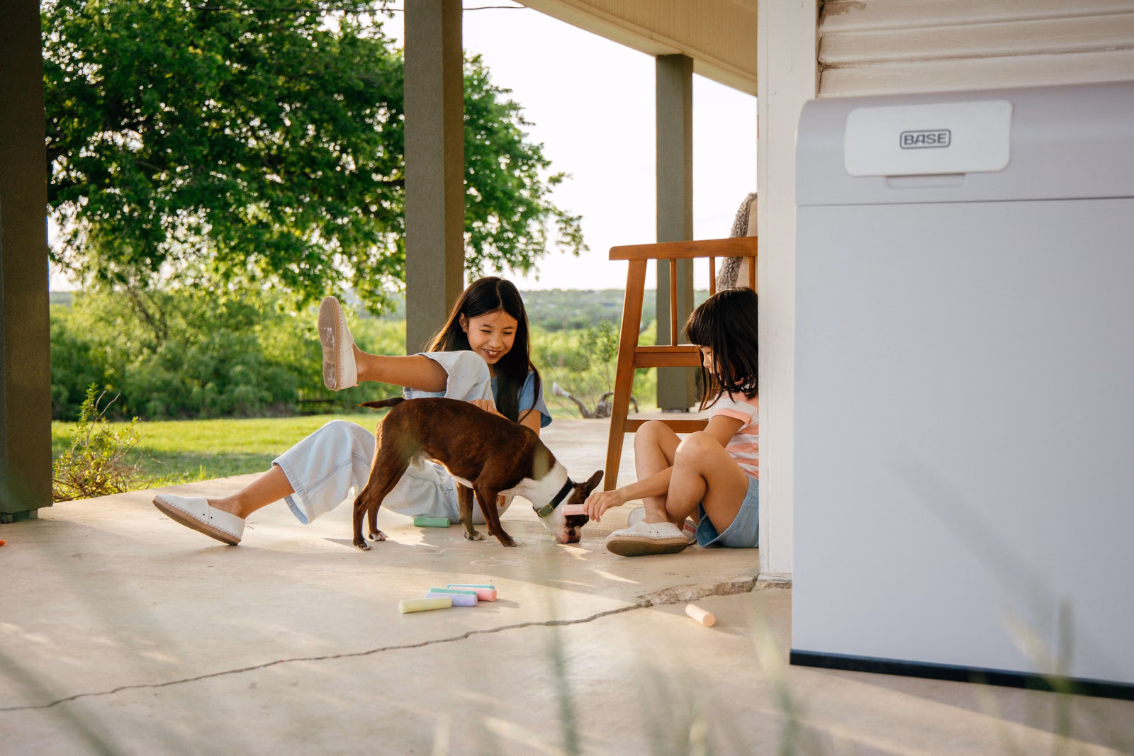 Children and a dog playing on a porch beside a Base Power battery