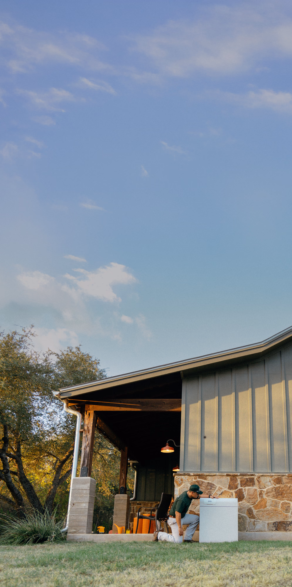 A Base technician kneeling beside a Base Power battery outside a stone-and-siding home at dusk