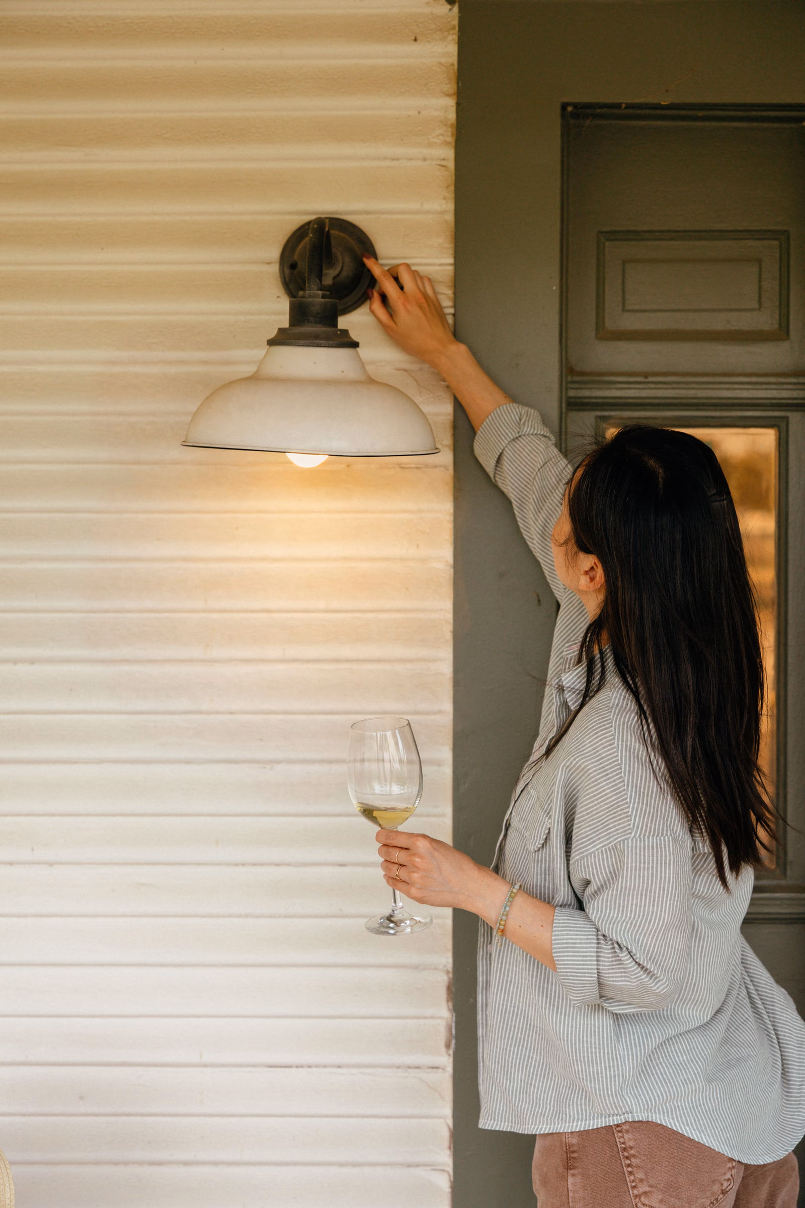 A woman turning on a porch light at home, holding a glass of wine